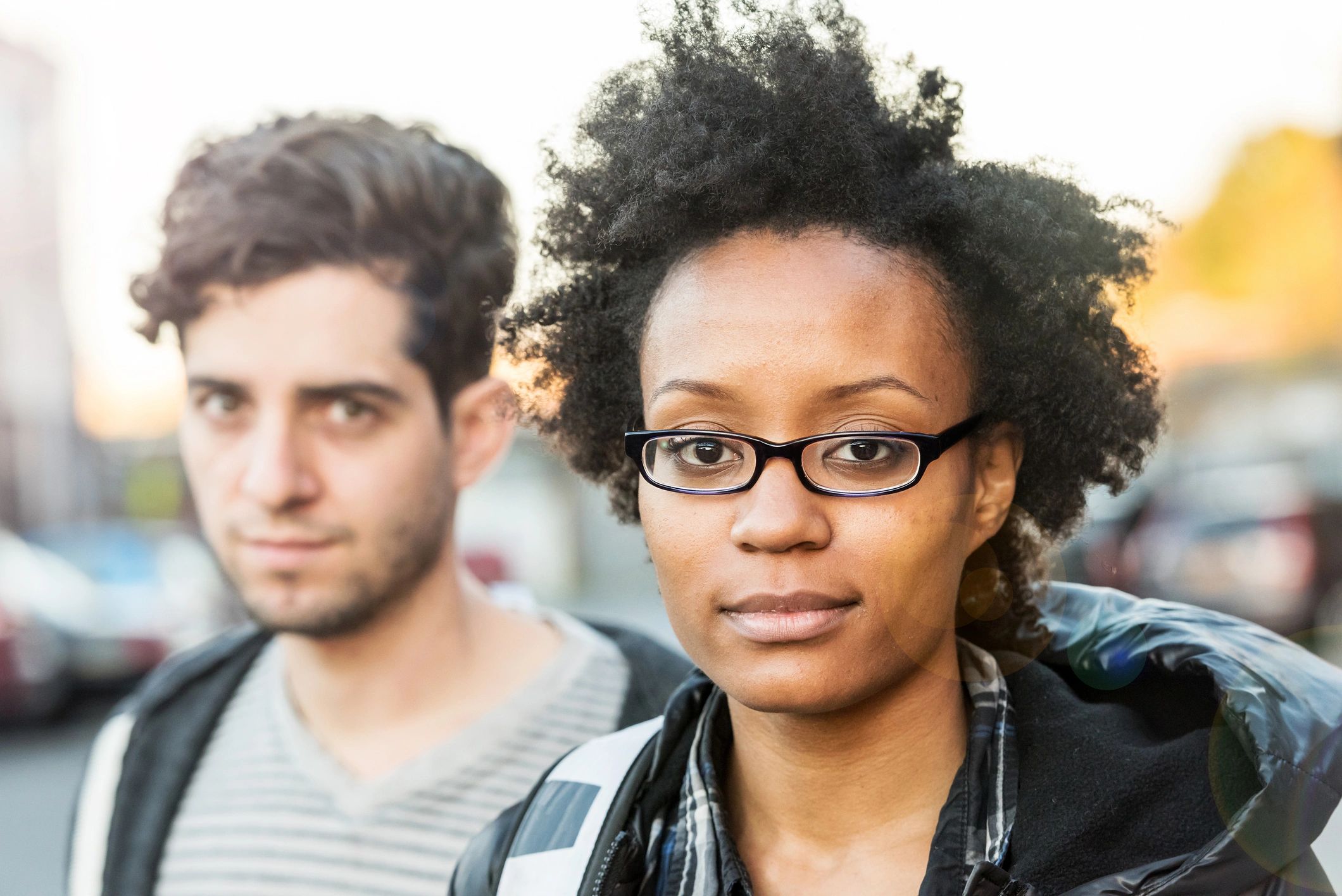 A young woman with glasses and curly hair stands outdoors with a blurred man in the background.